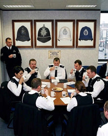 Police officers gathered around a table drinking tea beneath four framed artworks depicting police helmets.