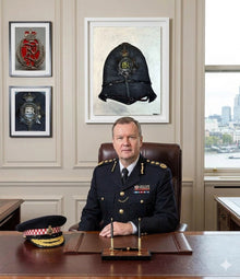 Uniformed police officer seated at a wooden desk beneath a framed police helmet artwork, with two framed badge artworks beside it.